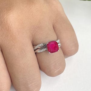 May include: A silver ring set featuring a large, square-cut, bright red gemstone. The band is adorned with small, sparkling accent stones. A second, matching band sits below the main ring. The jewelry is displayed on a light background.