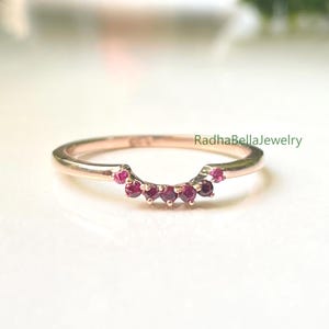 Op de afbeelding: Een roségouden ring met een gebogen ontwerp, bezet met kleine, levendige rode edelstenen. De ring wordt gepresenteerd op een witte ondergrond, met groene bladeren op de achtergrond. De tekst "RadhaBellaJewelry" is zichtbaar.