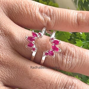 May include: A silver ring with a floral design, featuring vibrant pink gemstones. The ring is designed to wrap around another ring, creating a unique and eye-catching look. The gemstones are arranged in a petal-like pattern.