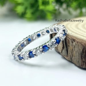 May include: A silver eternity ring with alternating blue and clear gemstones. The ring is set on a wooden surface with green foliage in the background. The text "RadhaBellaJewelry" is visible.