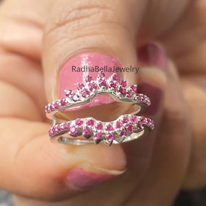 May include: Two silver-colored rings with a curved design, each adorned with numerous small, bright pink gemstones. The rings are held up against a blurred background. The text "RadhaBellaJewelry" is visible.