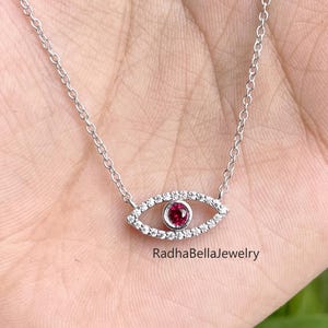 Op de afbeelding: Een zilveren ketting met een oogvormige hanger. De hanger heeft een centrale rode edelsteen omringd door kleine, heldere stenen. De ketting wordt getoond op een hand, met de merknaam "RadhaBellaJewelry".