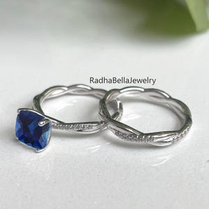 May include: A pair of silver rings. One ring features a square, deep blue gemstone. The other ring has a twisted design with small, clear stones. The rings are on a white surface. The text "RadhaBellaJewelry" is visible.