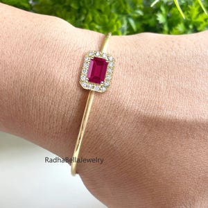May include: A gold bangle bracelet with a rectangular, deep red gemstone, encircled by smaller, clear stones. The minimalist design highlights the central gemstone. The text "RadhaBellaJewelry" is visible. The bracelet is a delicate piece of jewellery.