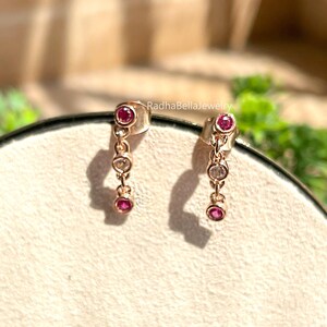 May include: A pair of rose gold dangle earrings. Each earring features three gemstones: a large, round, red gemstone at the top and bottom, and a smaller, clear gemstone in the middle. The earrings are displayed on a light beige surface.