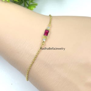 May include: A delicate gold bracelet featuring a rectangular ruby gemstone and two small diamonds. The bracelet is attached to a fine chain. The text "RadhaBellaJewelry" is visible on the bracelet.