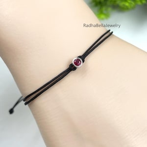 Peut inclure: Bracelet en fil noir avec une petite pierre précieuse rouge et ronde sertie dans un métal argenté. Le bracelet est noué avec une fermeture coulissante. La marque "RadhaBellaJewelry" est visible.