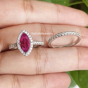 Op de afbeelding: Twee ringen, één met een marquise-geslepen robijn als middensteen, omgeven door een halo van kleine diamanten, en een tweede band met kleine diamanten. De ringen zijn gezet in een zilverkleurig metaal. De merknaam "RadhaBellaJewelry" is zichtbaar.