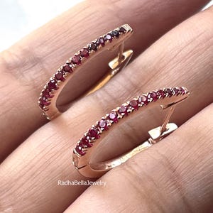 Könnte beinhalten: Ein Paar roségoldene Creolen, jeweils besetzt mit kleinen, leuchtend roten Edelsteinen. Die Ohrringe werden vor einem neutralen Hintergrund präsentiert, der ihr zartes Design und ihre funkelnden Details hervorhebt. Der Schmuck ist von RadhaBellaJewelry.