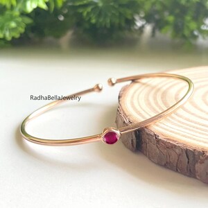 Könnte beinhalten: Ein offenes Armreif-Armband aus Roségold mit einem kleinen, runden, roten Edelstein. Das Armband wird auf einer Holzscheibe mit grünem Laub im Hintergrund präsentiert. Der Text "RadhaBellaJewelry" ist sichtbar.