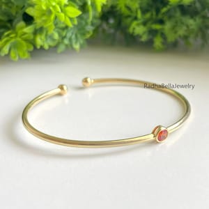 Puede incluir: Brazalete dorado con una pequeña gema redonda de color naranja. El brazalete tiene un diseño sencillo y abierto con extremos redondeados. El texto "RadhaBellaJewelry" es visible. Fondo de follaje verde.