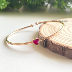 May include: A rose gold bangle bracelet with a heart-shaped, ruby-colored gemstone. The bracelet is displayed on a wooden slice with a green leafy background. The text "RadhaBellaJewelry" is visible.