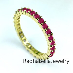 May include: A gold ring featuring a continuous line of small, bright red gemstones. The ring is set against a white backdrop with green foliage in the top right. The text "RadhaBellaJewelry" is visible on the ring.