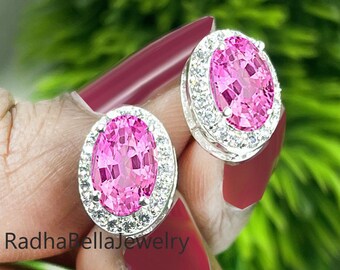 May include: A pair of oval-cut pink gemstone stud earrings with a halo of small, clear gemstones. The earrings are set in a silver-colored metal. The background is blurred with green foliage.