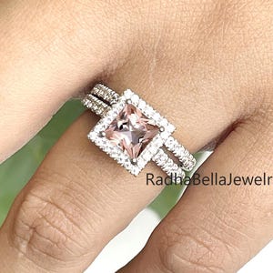 Puede incluir: Un anillo de piedra preciosa rosa claro de corte cuadrado con un halo de pequeñas piedras transparentes. El anillo está engarzado en una banda plateada con piedras pequeñas adicionales. El texto "RadhaBellaJewelry" es visible.
