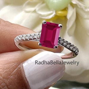 May include: A silver ring featuring a large, rectangular, ruby-red gemstone. The band is embellished with smaller, clear gemstones. The ring is held against a blurred floral backdrop. The text "RadhaBellaJewelry" is visible.
