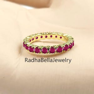 May include: A gold-colored eternity ring with a band of vibrant red gemstones. The ring is set against a neutral background, with the text "RadhaBellaJewelry" visible. The ring is a piece of jewelry.