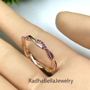 以下が含まれることがあります： ピンク色の宝石が埋め込まれた、ひねりのあるデザインのローズゴールドリング。リングはニュートラルな背景に配置され、その複雑なディテールが際立っています。「RadhaBellaJewelry」の文字がリングに表示されています。