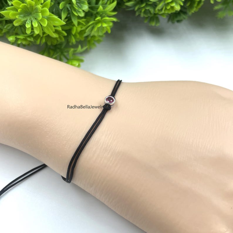 May include: A delicate black string bracelet featuring a small, round, purple gemstone set in silver-coloured metal. The bracelet is displayed on a wrist, highlighting its simple design. The brand name "RadhaBellaJewels" is visible.