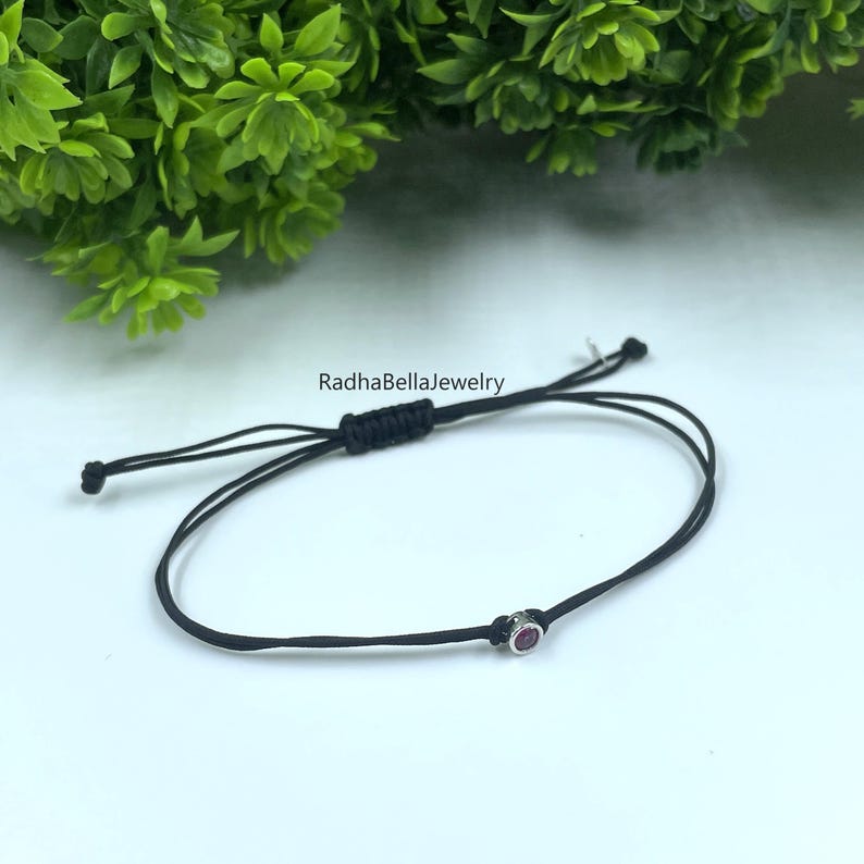 May include: Black adjustable bracelet with a small, round, pink gemstone. The bracelet is made of a thin, black cord and features a sliding knot for adjustment. The background includes green foliage and the text "RadhaBellaJewelry".