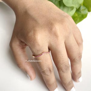 Peut inclure: Une bague dorée avec un design incurvé, sertie de petites pierres précieuses rouges vibrantes. La bague est portée au doigt, mettant en valeur le savoir-faire délicat et l'éclat des pierres. La bague est de RadhaBella Jewelry.