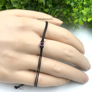 May include: A fine black cord bracelet with a small, round, ruby-coloured gemstone set in a silver bezel. The bracelet is displayed on a hand, with the brand name "RadhaBellaJewelry" visible in the background.