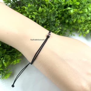 May include: A black string bracelet featuring a small, oval, dark pink gemstone. The bracelet is on a wrist, with the text "RadhaBellaJewelry" visible. Green foliage forms the background.