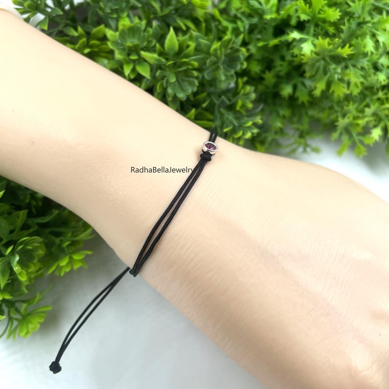 May include: A black string bracelet featuring a small, oval, dark pink gemstone. The bracelet is on a wrist, with the text "RadhaBellaJewelry" visible. Green foliage forms the background.