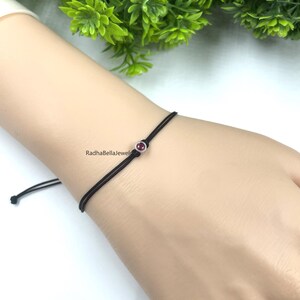 May include: A black cord bracelet featuring a small, round, red gemstone set in a silver bezel. The bracelet is on a wrist, with the brand name "RadhaBellaJewel" visible. Green foliage is in the background.