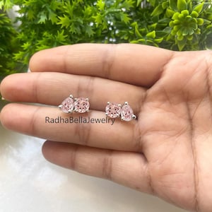 May include: A pair of stud earrings featuring a round and a pear-shaped pink gemstone. The earrings are set in silver and are displayed on a hand. The text "RadhaBellaJewelry" is visible.