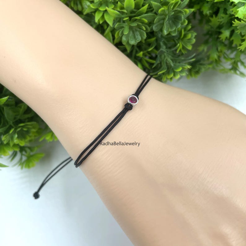 May include: Black string bracelet featuring a small, round, red gemstone set in silver-coloured metal. The bracelet has an adjustable sliding knot. The text "RadhaBellaJewelry" is printed on the arm.