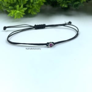 May include: Black cord bracelet with a small, round, silver-toned charm featuring a vibrant pink gemstone. The bracelet has an adjustable sliding knot closure. The text "RadhaBellaJewelry" is visible on the bracelet.