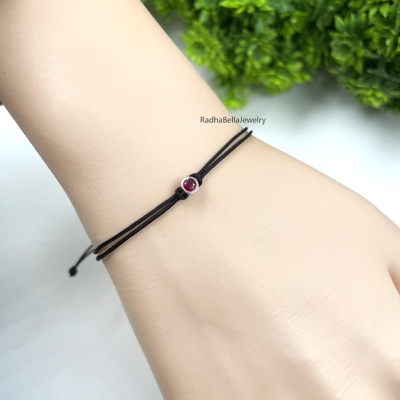 May include: Black string bracelet featuring a small, round, red gemstone set in a silver-coloured metal. The bracelet has a sliding knot closure. The brand name "RadhaBellaJewelry" is visible in the background.