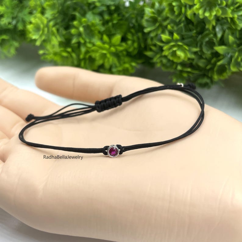 May include: Black cord bracelet featuring a small, round, ruby-coloured gemstone set in a silver-coloured bezel. The bracelet is knotted for adjustability. The text "RadhaBellaJewelry" is visible on the bracelet.