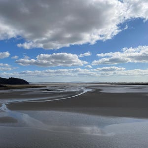 May include: A scenic landscape featuring a wide expanse of wet sand and shallow water under a partly cloudy sky. The sky is a bright blue with fluffy white clouds. A dark, tree-covered hill is visible in the distance.