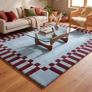 May include: A rectangular area rug featuring a light blue centre and a dark red geometric border pattern consisting of repeating rectangular blocks. The rug is laid flat on a wooden floor, showcasing a textured pile surface and clean edges.