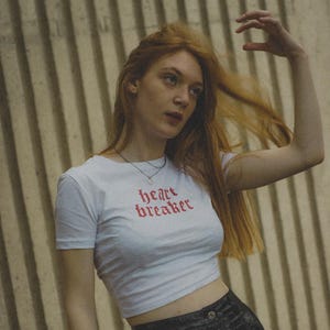 Op de afbeelding: Een witte crop top met de tekst "heart breaker" in rode letters. De top wordt gecombineerd met een zwarte leren rok met een grote zilveren ring en knopen. De persoon heeft lang rood haar.