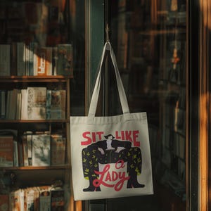 May include: Cream-colored tote bag with the text "SIT LIKE A LADY" in red and black. The bag features an illustration of a person sitting in a chair with a black jacket and patterned pants. The bag is hanging on a hook.