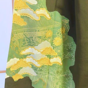 May include: Close-up of a fabric with a Japanese-inspired design. The fabric features a green background with yellow and white cloud and building patterns. The edges are a darker green, creating a layered effect. The design is detailed and intricate.