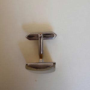 May include: A single silver-toned cufflink with a rectangular bar and a rounded, oval-shaped end. The cufflink has a T-shaped design, with a central post connecting the two ends. The metal has a slightly tarnished appearance.
