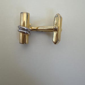 May include: A single gold-toned cufflink with a rectangular face and a cylindrical post. The post is wrapped with silver-colored metal. The cufflink is designed to fasten a shirt cuff.