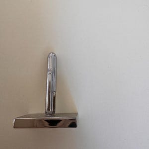 May include: A small, shiny, silver-colored metal object with a rectangular base and a cylindrical top. The object is standing upright against a plain, off-white background. The object appears to be a decorative item or a small sculpture.