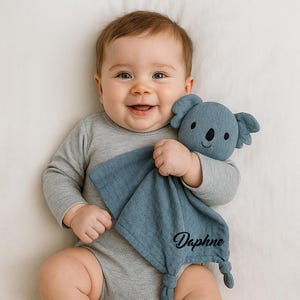 Pu&ograve; includere: Un bambino sorridente in un body grigio tiene in mano una coperta di sicurezza a forma di koala blu. La coperta ha il nome "Daphne" ricamato in nero. Il koala ha un naso nero e una faccia sorridente.