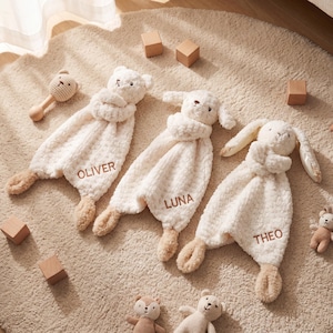 May include: Three plush baby security blankets with animal heads, each personalized with a name: "OLIVER", "LUNA", and "THEO". The blankets are cream-colored with tan accents and are surrounded by wooden blocks and other stuffed toys.