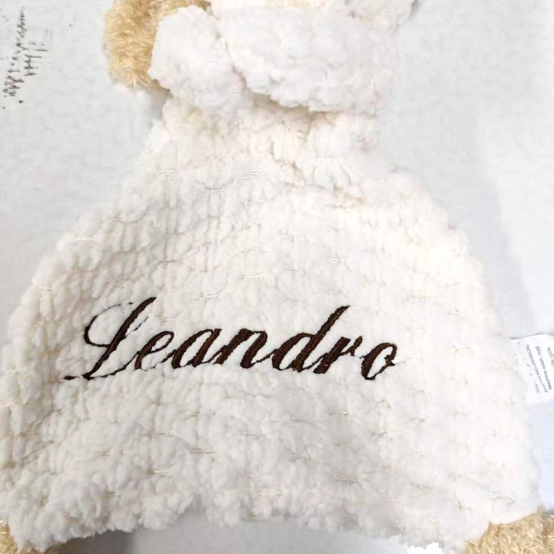 May include: A plush, cream-colored baby blanket with the name "Leandro" embroidered in brown script. The blanket has a textured, fluffy surface and a light tan trim.