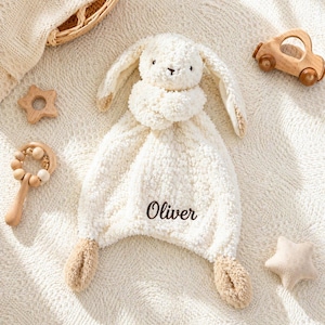May include: A cream-colored baby security blanket shaped like a bunny with the name "Oliver" embroidered on it. The blanket is surrounded by wooden toys, including a star, a rattle, and a car.