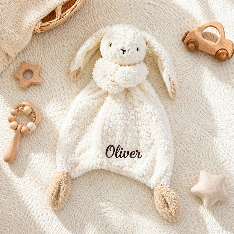 May include: A cream-colored baby security blanket shaped like a bunny with the name "Oliver" embroidered on it. The blanket is surrounded by wooden toys, including a star, a rattle, and a car.