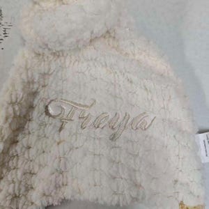 May include: Cream-colored, plush baby hat with the name "Freya" embroidered in a delicate script. The hat has a soft, textured appearance and a rounded top. The fabric appears to be a cozy, warm material.