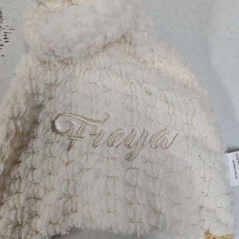 May include: Cream-colored, plush baby hat with the name "Freya" embroidered in a delicate script. The hat has a soft, textured appearance and a rounded top. The fabric appears to be a cozy, warm material.