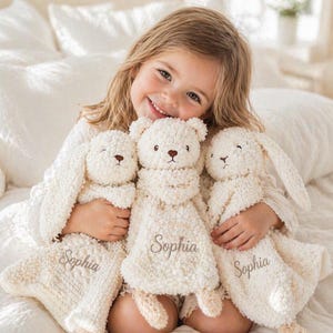May include: A young girl smiles while holding three cream-colored plush toys: a bear and two bunnies. Each toy has a brown nose and the name "Sophia" embroidered on it. The toys are soft and fluffy, perfect for a child.